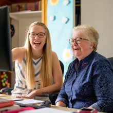 A woman and an older woman seated at a desk, engaged in conversation while using a computer.