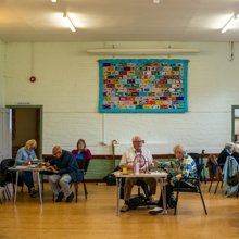 Group of senior citizens participating in a game , seated around tables in a community hall, with a large patchwork quilt on the wall.