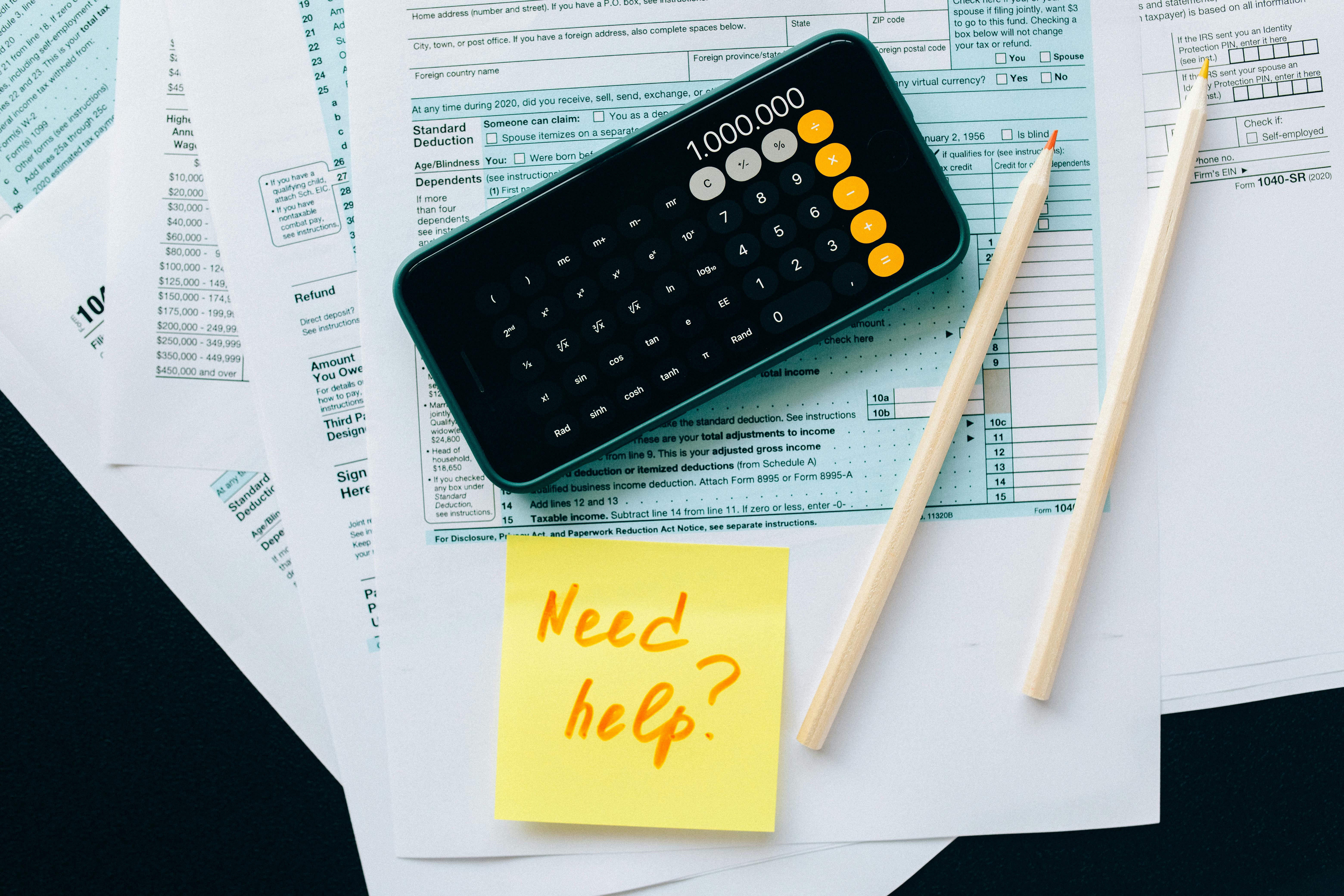 Official forms, calculator, and a sticky note reading "Need help?" are laid out on a dark surface.