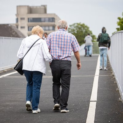 Older couple walking arm-in-arm on a paved walkway, with a metal fence in the background.